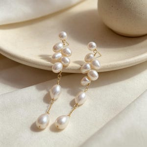 May include: A pair of drop earrings featuring multiple white freshwater pearls arranged in a cascading cluster design. The pearls are connected by thin gold-toned metal chains and links, with a single pearl stud at the top of each earring.