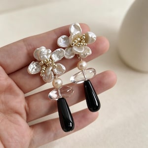 Può includere: Un paio di orecchini eleganti con grappoli di perle a forma di fiore con dettagli dorati. Ogni orecchino ha una perla, un cristallo trasparente e una pietra nera a forma di goccia, creando un accessorio sofisticato ed elegante.