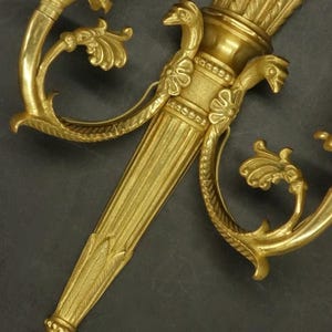 Puede incluir: Un aplique de pared de metal dorado con un diseño central en forma de antorcha, un eje estriado y un haz de flechas en la parte superior. Dos brazos curvos se extienden desde los lados, terminando en portavelas con fundas blancas.