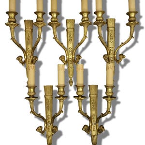 May include: A set of five gold-toned metal wall sconces, each featuring multiple candle-style light fixtures. The sconces have ornate vertical backplates with textured detailing, curved arms, and decorative finials at the base of each unit.