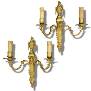 May include: A pair of gold-toned metal wall sconces, each featuring two candle-style light fixtures with cream-colored drip-effect sleeves. The central vertical support is adorned with an urn-shaped finial and ornate relief details.