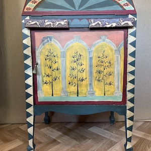 May include: A hand-painted wooden cabinet in teal, red, and yellow. The cabinet has an arched design with olive trees and a starburst pattern. The legs are painted with a white and teal triangle pattern. The cabinet measures approximately 150 cm in height.