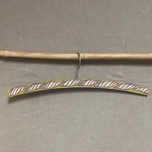 May include: A decorative wooden clothes hanger with a green base and a purple and white wave pattern. The hanger is suspended from a silver metal hook and a bamboo rod.