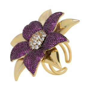 Op de afbeelding: Een goudkleurige bloemenring met een opvallend ontwerp. De bloemblaadjes zijn versierd met kleine, dieppaarse edelstenen, en het midden heeft heldere, sprankelende stenen. De ring loopt rond de vinger.