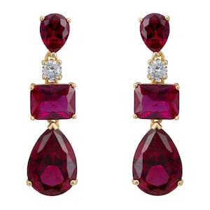 以下が含まれることがあります： 3つのルビーレッドの宝石が付いた、ゴールドトーンのドロップイヤリングのペア。上部の石は小さな楕円形で、四角い石、大きな涙型の石が続きます。上部と中央の石の間には、小さな透明な石があります。