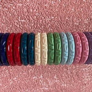 Puede incluir: Un conjunto de 12 pulseras de plástico de colores con un diseño floral tallado. Las pulseras están dispuestas en un patrón de arcoíris, comenzando con azul oscuro y terminando con morado.
