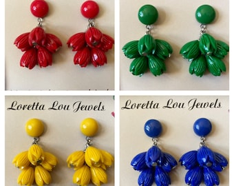 May include: Four pairs of Loretta Lou Jewels earrings in red, green, yellow, and blue. Each pair features a round stud and a cluster of flower-shaped dangles. The earrings are displayed on a cream-colored background.