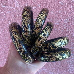 May include: A set of five black bangle bracelets with gold glitter accents. The bracelets are stacked on top of each other.