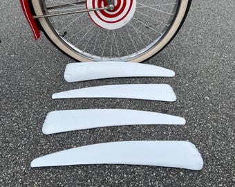 May include: Four white, curved, elongated plastic bicycle wheel spoke covers arranged horizontally. The covers feature a smooth, tapered design with a rounded end and a slightly wider, notched base, positioned in front of a bicycle wheel with a red and white target-patterned disc.
