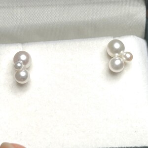 Peut inclure: Une paire de boucles d'oreilles en grappe de perles présentée dans un écrin à bijoux noir avec un intérieur blanc. Chaque boucle d'oreille comporte trois ou quatre perles blanches et blanc cassé, créant un design délicat. Les perles ont un éclat brillant.