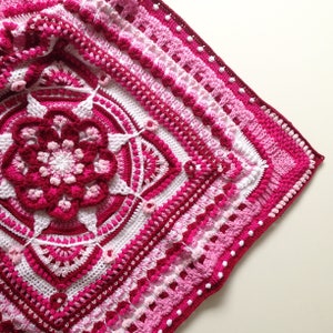 Puede incluir: Una manta de crochet rosa y blanca con un gran diseño de flor en el centro. La manta está hecha con una variedad de puntos y texturas.