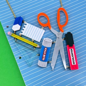 Könnte beinhalten: Ein bunter Satz an Schulbedarfs-Anhängern, darunter ein blauer Radiergummi, ein weißer Notizblock, ein gelber Bleistift, ein Klebestift, eine orangefarbene Schere und ein rosa Textmarker. Die Anhänger sind an einer silbernen Kette befestigt.