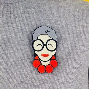 以下が含まれることがあります： グレーと白の漫画の肖像画のブローチで、赤い丸いビーズが付いています。ブローチは、灰色の髪、黒い眼鏡、赤い唇をした女性を描いています。