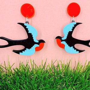 Könnte beinhalten: Ein Paar Ohrringe mit einem schwarzen und blauen Schwalbenmotiv mit einem roten Akzent. Die Ohrringe hängen an einem roten Kreis.