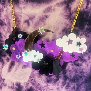 May include: A gold chain necklace with a purple, black and white crescent moon and cloud pendant. The pendant features iridescent stars and a reflective surface.