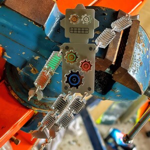 Puede incluir: Un amuleto de robot plateado con engranajes coloridos y una cadena plateada. El robot sostiene una cadena plateada con un amuleto plateado y verde.