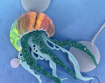 May include: A teal and orange glitter jellyfish pendant with a blue and green iridescent finish. The pendant is attached to a silver chain with a clear plastic disc.