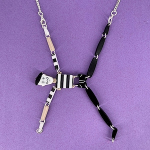 May include: A black and white striped figure necklace with a silver chain. The figure has a black beret and the text '+3' on its face.