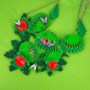 Peut inclure: Un collier vert et rouge avec un motif floral. Le collier est en plastique et présente une variété de fleurs et de feuilles. Le collier est une pièce forte et serait parfait pour une fête d'été ou une soirée.