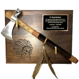 May include: A wooden plaque with a tomahawk and peace pipe mounted on it. The plaque has an engraved image of an American Indian chief with a skull. The plaque also has a brass plate with the text "M. Greg Ramirez We express our deep appreciation for your outstanding dedication and service in support of the Waco Recruiting Company 15 SEP 2020 - 37 OCT 2026 On behalf of the Waco Warrior Soldiers and Command".