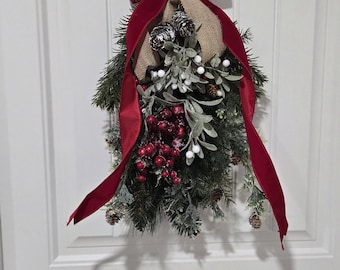 May include: A Christmas swag with red velvet ribbon, pine sprigs, berries, and pinecones hanging on a white door.