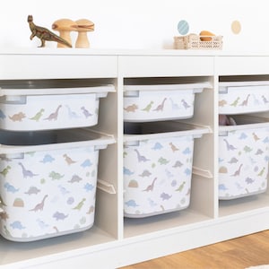 Può includere: Scaffali di stoccaggio bianchi con quattro contenitori di plastica. I contenitori hanno uno sfondo bianco con un motivo di dinosauri colorati.