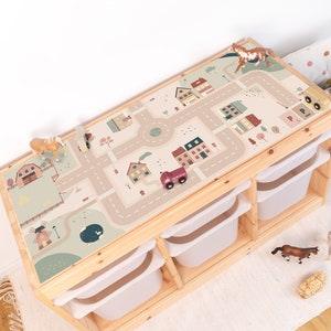 Könnte beinhalten: Ein Holzspieltisch mit einer abnehmbaren Spielmatte, die eine Stadtlandschaft mit Häusern, Straßen und Bäumen zeigt. Die Spielmatte ist beige mit braunen, roten und grünen Akzenten. Der Tisch hat vier Aufbewahrungskisten.
