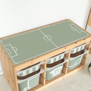 Peut inclure: Un rangement de jouets en bois avec quatre bacs rayés vert et blanc. Le dessus de l'unité a une surface verte avec un motif de terrain de football blanc. Un bac a un maillot de football blanc avec le nom "Moritz" et le numéro "11" imprimé dessus.