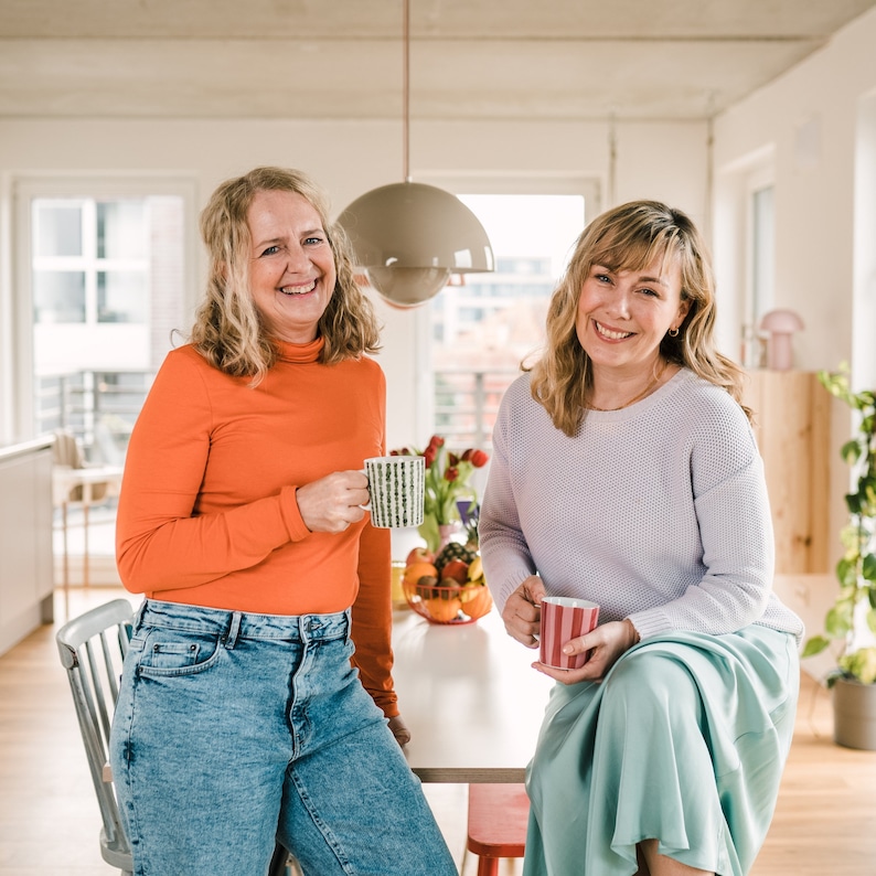 May include: Two women are smiling and holding mugs. The woman on the left is wearing a bright orange turtleneck sweater and blue jeans. The woman on the right is wearing a lavender sweater and a light blue skirt. They are sitting at a table with a white tablecloth and a bowl of fruit.