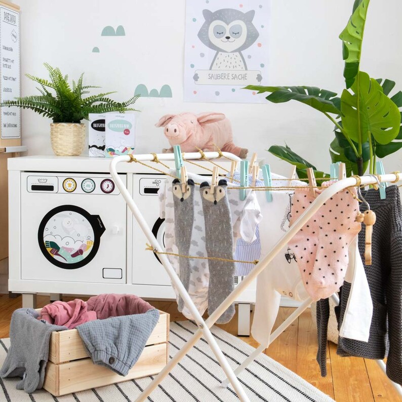 May include: White metal drying rack with clothes hanging on it. The rack is in a room with a white washing machine and a wooden crate. The clothes are a variety of colors and patterns, including white, gray, pink, and blue. The text "Saubere Sache" is visible on a poster in the background.