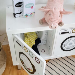 May include: White toy washing machine with two doors, one open to reveal a yellow and blue shirt inside. The washing machine has a black and white design with a dial and buttons. A pink stuffed pig is on top of the machine.