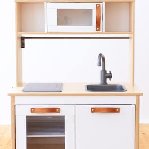 Op de afbeelding: Witte houten speelkeuken met een zwart fornuis, een grijze gootsteen en een witte magnetron. De keuken heeft bruine leren handvatten en een metalen stang om keukengerei aan op te hangen.