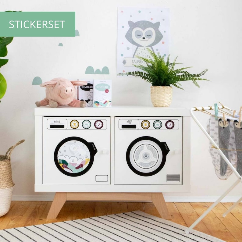 May include: A white toy washing machine set with two washing machines and a drying rack. The washing machines have black knobs and a black circle in the center of the door. The drying rack has a gray and white patterned cloth hanging on it. The set is on a wooden floor with a striped rug in front of it.