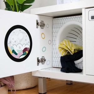 May include: White toy washing machine with a door that opens to reveal a colorful cartoon design and a white laundry basket. The machine has a white base with silver legs.