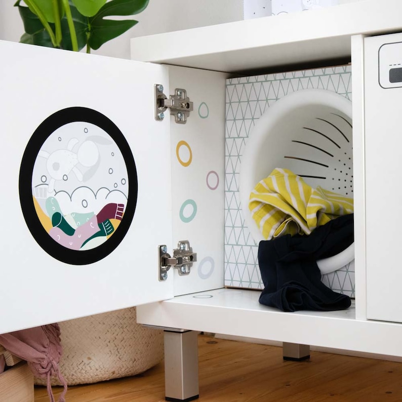 May include: White toy washing machine with a door that opens to reveal a colorful cartoon design and a white laundry basket. The machine has a white base with silver legs.
