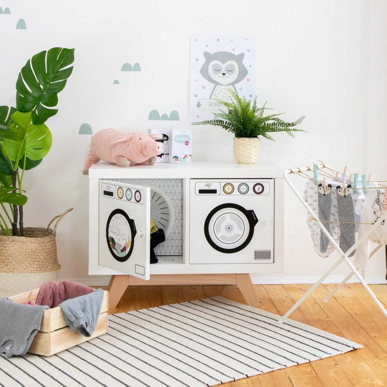 May include: A white toy washing machine with two doors, one open to reveal a spinning drum. The machine is on a wooden stand with a pink stuffed pig and a green plant in the background.