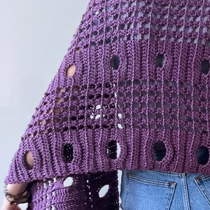 May include: A purple crochet shawl with large circular cutouts. The shawl is made of a textured yarn and has a loose, open weave design. The garment is worn over a gray top and blue jeans.