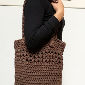 May include: A brown crocheted tote bag with a wide, open top and a woven pattern. The bag is being held by a person wearing a black long-sleeved shirt.