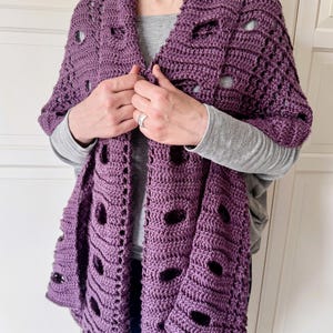 Könnte beinhalten: Ein gehäkelter lila Schal mit großen, gleichmäßig verteilten Öffnungen. Der Schal ist über die Schultern einer Person drapiert, wobei die Kanten herunterhängen. Das Garn scheint ein weiches, strukturiertes Material zu sein. Der Schal ist einfarbig, ohne weitere sichtbare Muster.