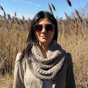 Puede incluir: Una mujer con una bufanda infinita de punto marrón y gafas de sol frente a un campo de juncos marrones altos.