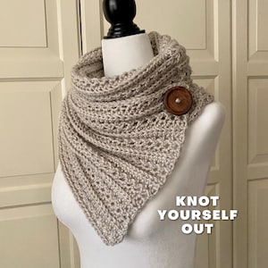 Puede incluir: Una bufanda infinita de crochet beige con un botón de madera. La bufanda está drapeada sobre un maniquí blanco. El texto "Knot Yourself Out" está impreso debajo de la bufanda.