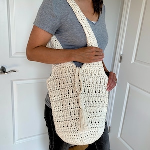 May include: A white crocheted tote bag with a long strap and a tassel. The bag is worn over the shoulder of a person wearing a gray shirt and jeans.