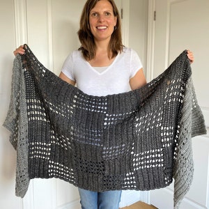 May include: A grey and white crocheted shawl with a lacy pattern. The shawl is draped over the shoulders of a person wearing a white shirt and blue jeans.