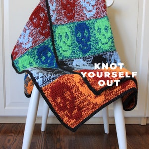 May include: A colorful crocheted blanket with a black border, featuring a skull pattern in green and blue squares. The blanket is draped over a white chair with the text "Knot Yourself Out" visible.