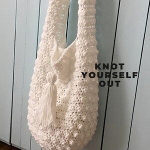 May include: White crocheted tote bag with a tassel hanging from the bottom. The bag is hanging on a light blue wooden wall. The text "KNOT YOURSELF OUT" is printed on the wall.