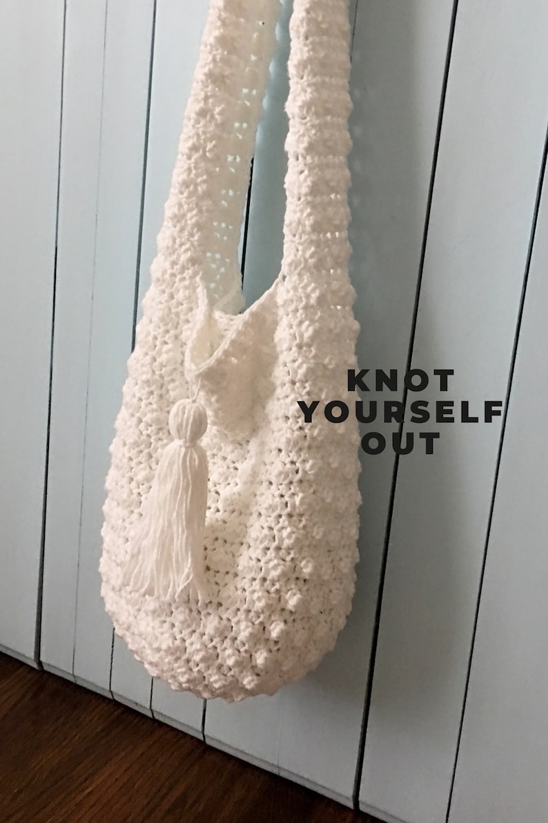 May include: White crocheted tote bag with a tassel hanging from the bottom. The bag is hanging on a light blue wooden wall. The text "KNOT YOURSELF OUT" is printed on the wall.