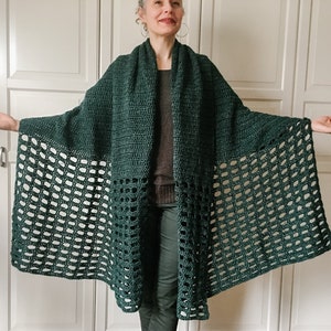 May include: A dark green crochet shawl with a lacy pattern. The shawl is draped over a person's shoulders and is worn with a dark brown sweater and dark green pants.
