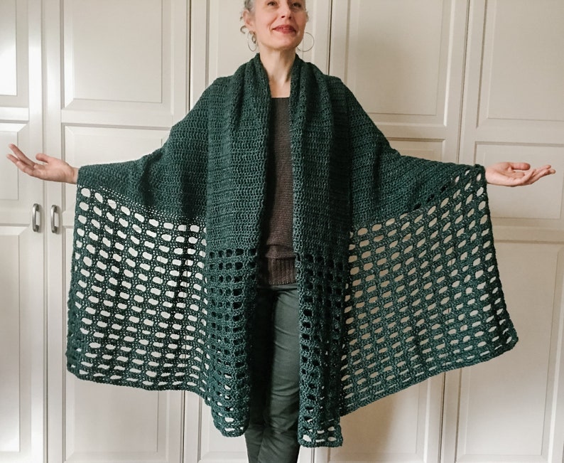 May include: A dark green crochet shawl with a lacy pattern. The shawl is draped over a person's shoulders and is worn with a dark brown sweater and dark green pants.