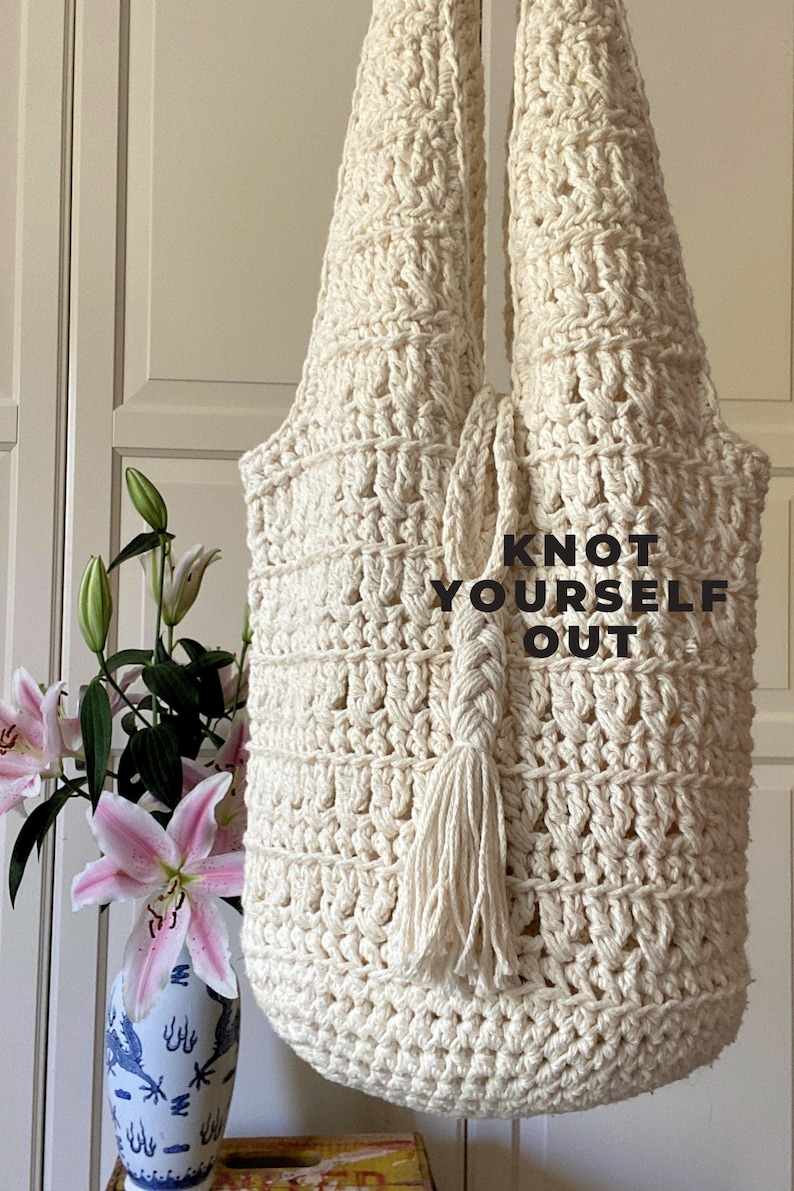May include: A cream-colored crocheted tote bag with a tassel. The bag has a knotted strap and the text "Knot Yourself Out" is printed on the front.