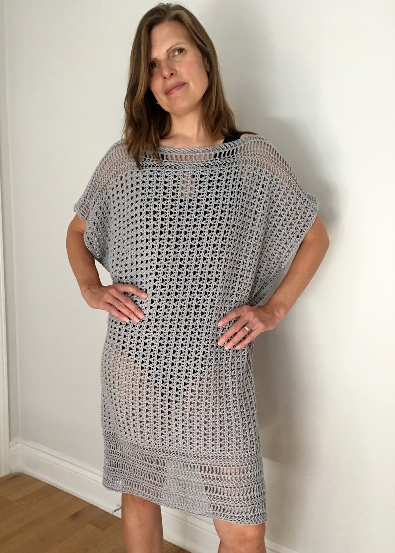Puede incluir: Un vestido de crochet gris de corte holgado y escote amplio. El vestido tiene un patr&oacute;n simple de tejido abierto y una falda ligeramente acampanada.